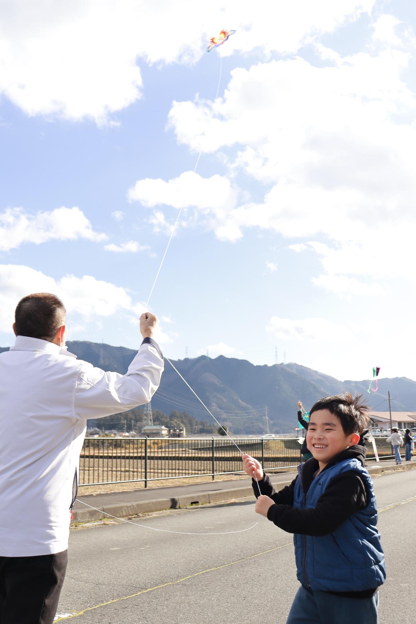 空高く揚がった凧が飛んでいかないように大人がたこ糸を抑え、男の子が糸の先をもって走り出そうとしている写真