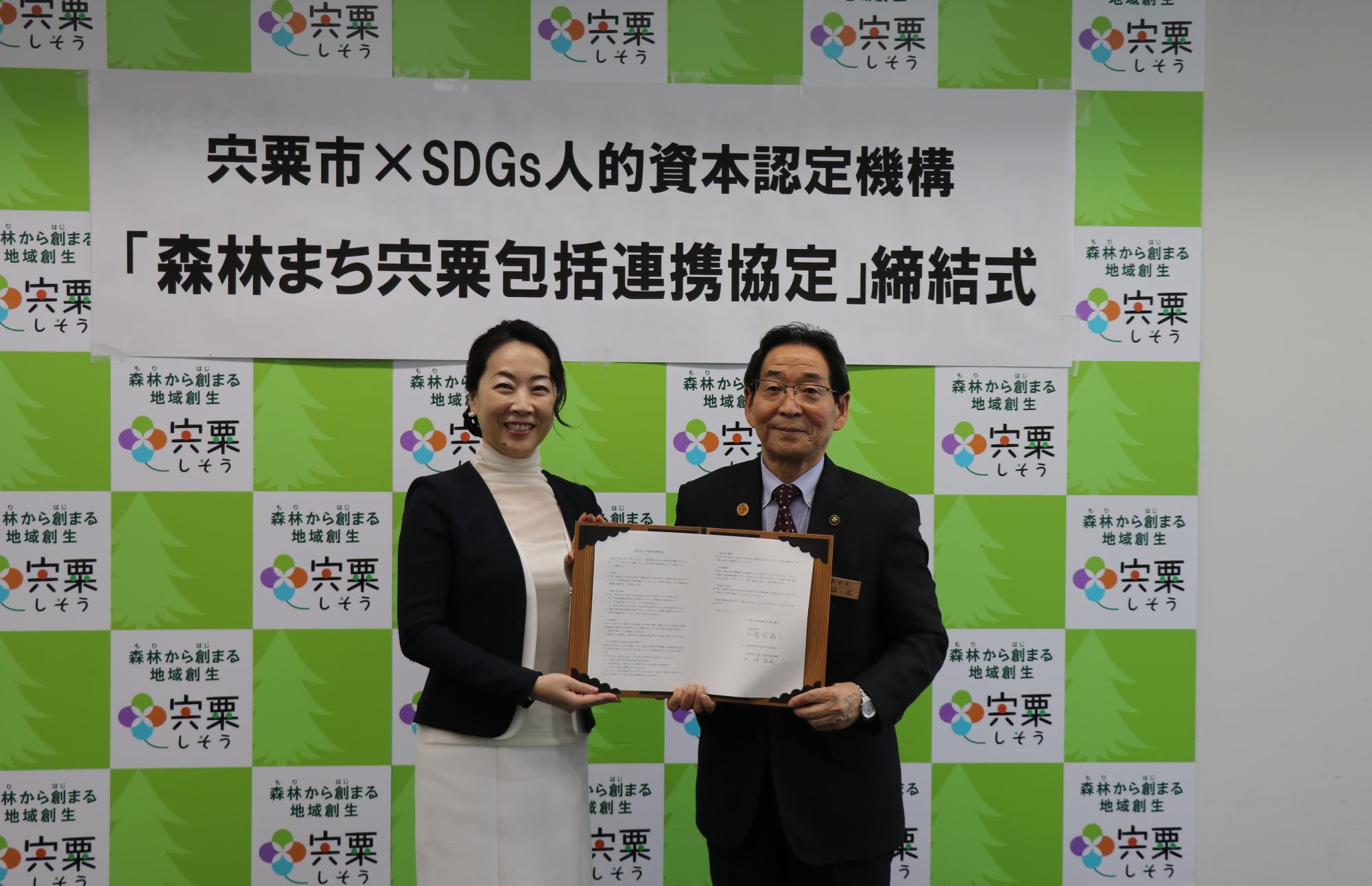森林まち宍粟包括連携協定を締結したSDGs人的資本認定機構の伴 裕美理事と福元晶三宍粟市長が横に並んで協定書を一緒に持ちほほえんでいる写真、
