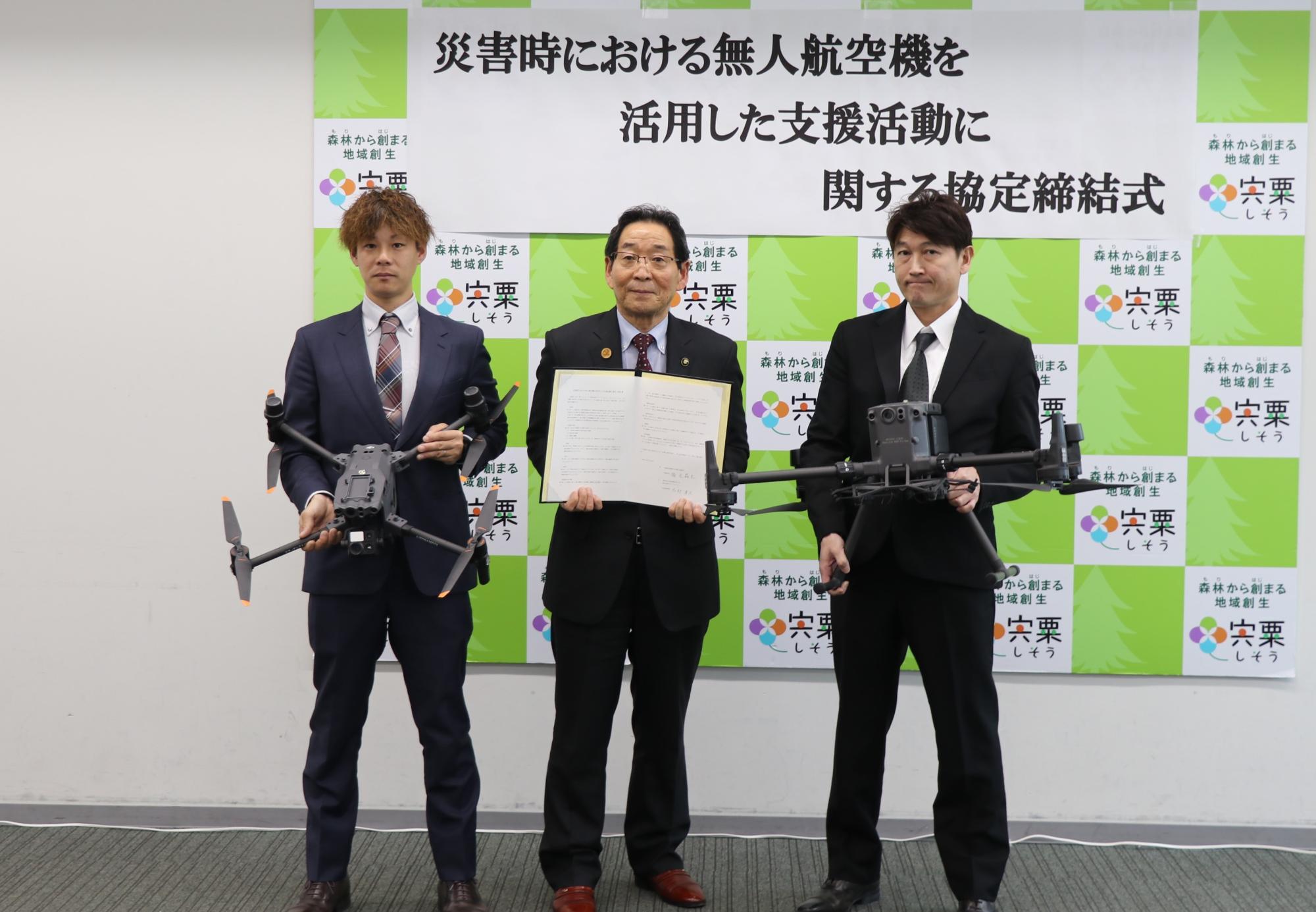 災害時における無人航空機を活用した支援活動に関する協定をそれぞれ締結した株式会社エアーアシストジャパン椿祐樹代表取締役と福元晶三宍粟市長と株式会社ドリームクエスト志村孝夫代表取締役の三人が協定書とドローンを手に持ち横に並んでいる写真