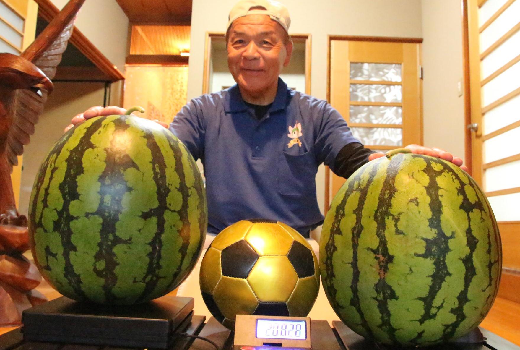 仲川光弘さんが向かって左に20キロ超のスイカを、右に16キロ超のスイカを、真ん中に大きさを比較するためにサッカーボールを置き、仲川さんがその後ろに座ってスイカに手を添え笑っている写真