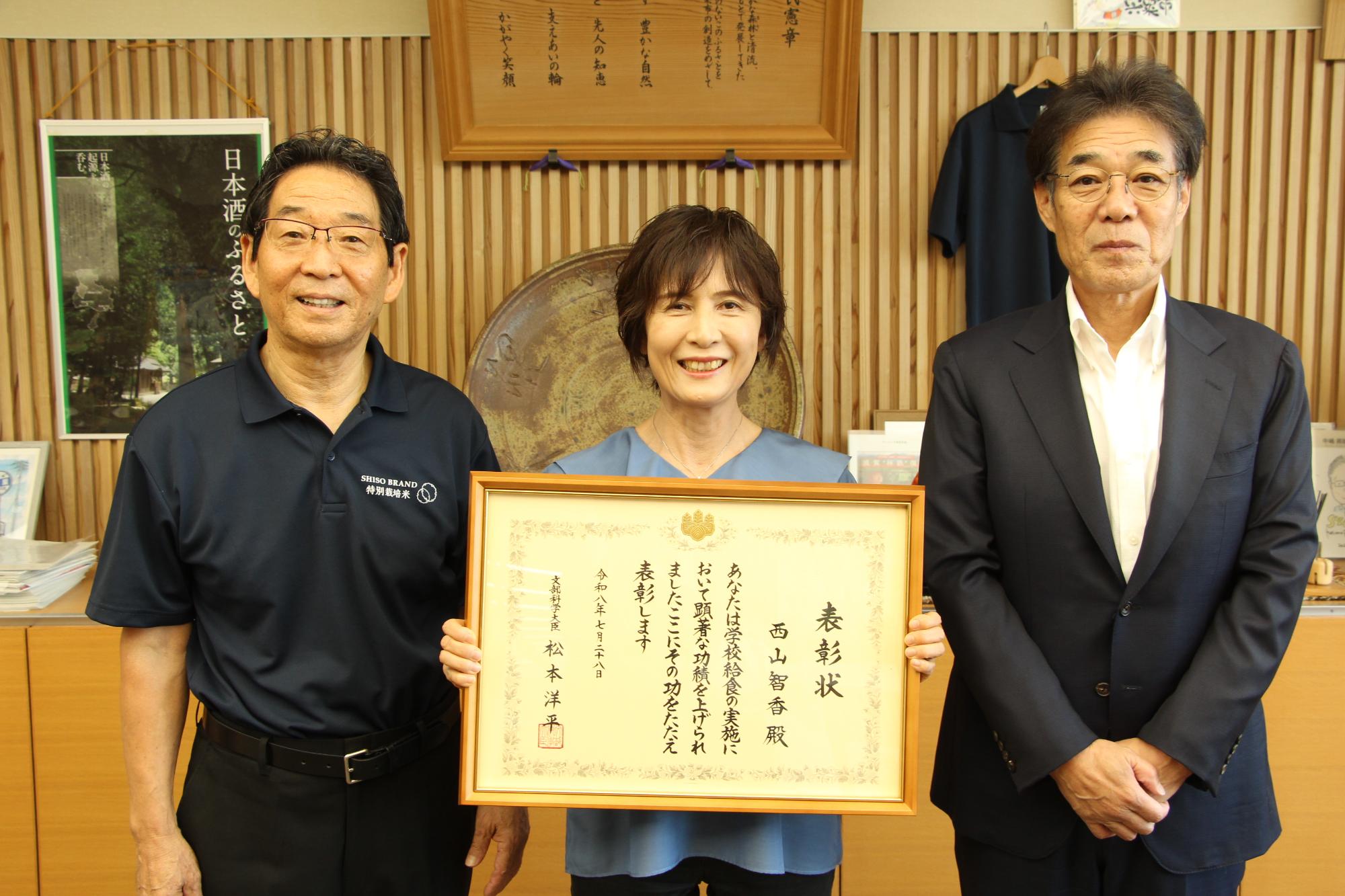 表彰状を抱える西山さんと、両脇に立つ福元市長と中田教育長