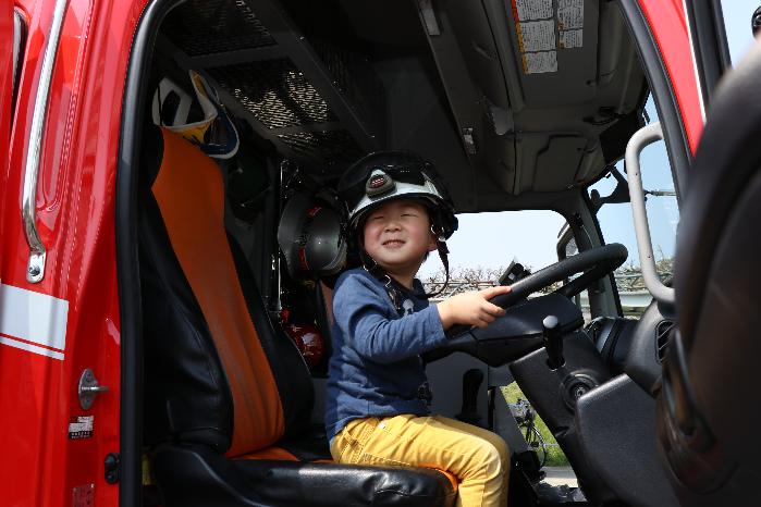 黒いヘルメットをかぶった男子小学生が消防車の運転席でハンドルをにぎっている写真
