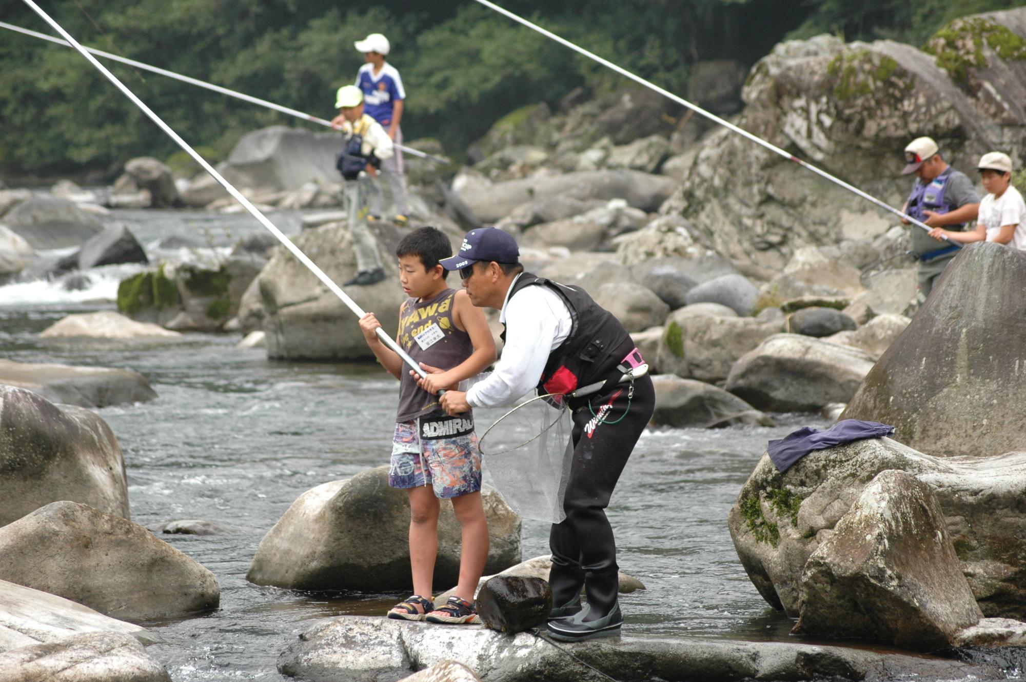 川で釣り客に鮎釣りを教えてもらっている子どもと釣り客の写真