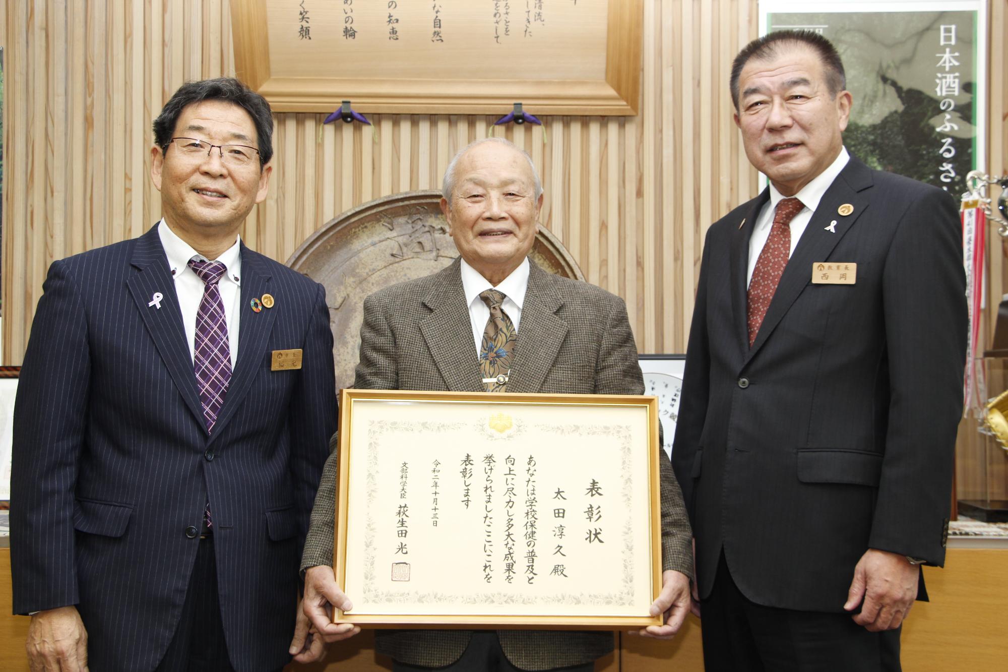 文部科学大臣表彰の額を手にした太田医師とそれを囲む福元市長と西岡教育長の写真