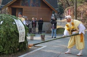 山伏の格好の人が人の背丈ほどある緑色の塊に松明を使い着火しようとしている写真