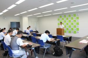 懇談会に出席した記者達と演壇に立つ白いシャツを着た男性の写真