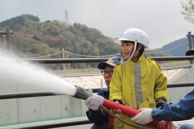 ヘルメットと消防服を着て水が出ている放水ホースを大人と一緒に持つ子どもの写真