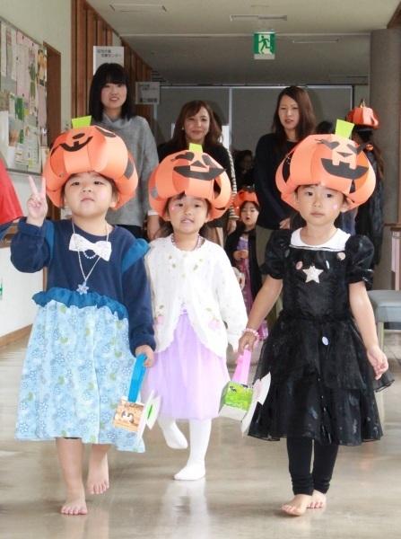 オレンジの紙で作られたかぼちゃ帽子をかぶって並んで廊下を歩く三人の子どもたちの写真