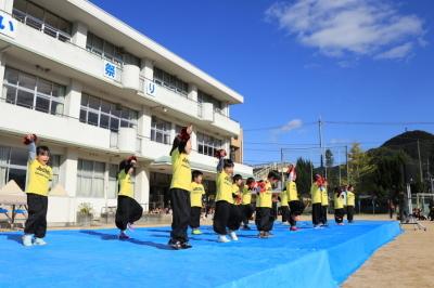 城下ふれあいまつりの小学校グラウンドに設置された水色のステージじょうでおそろいの黄色いTシャツと黒いズボンの子どもたちが右手お上げて踊っている写真