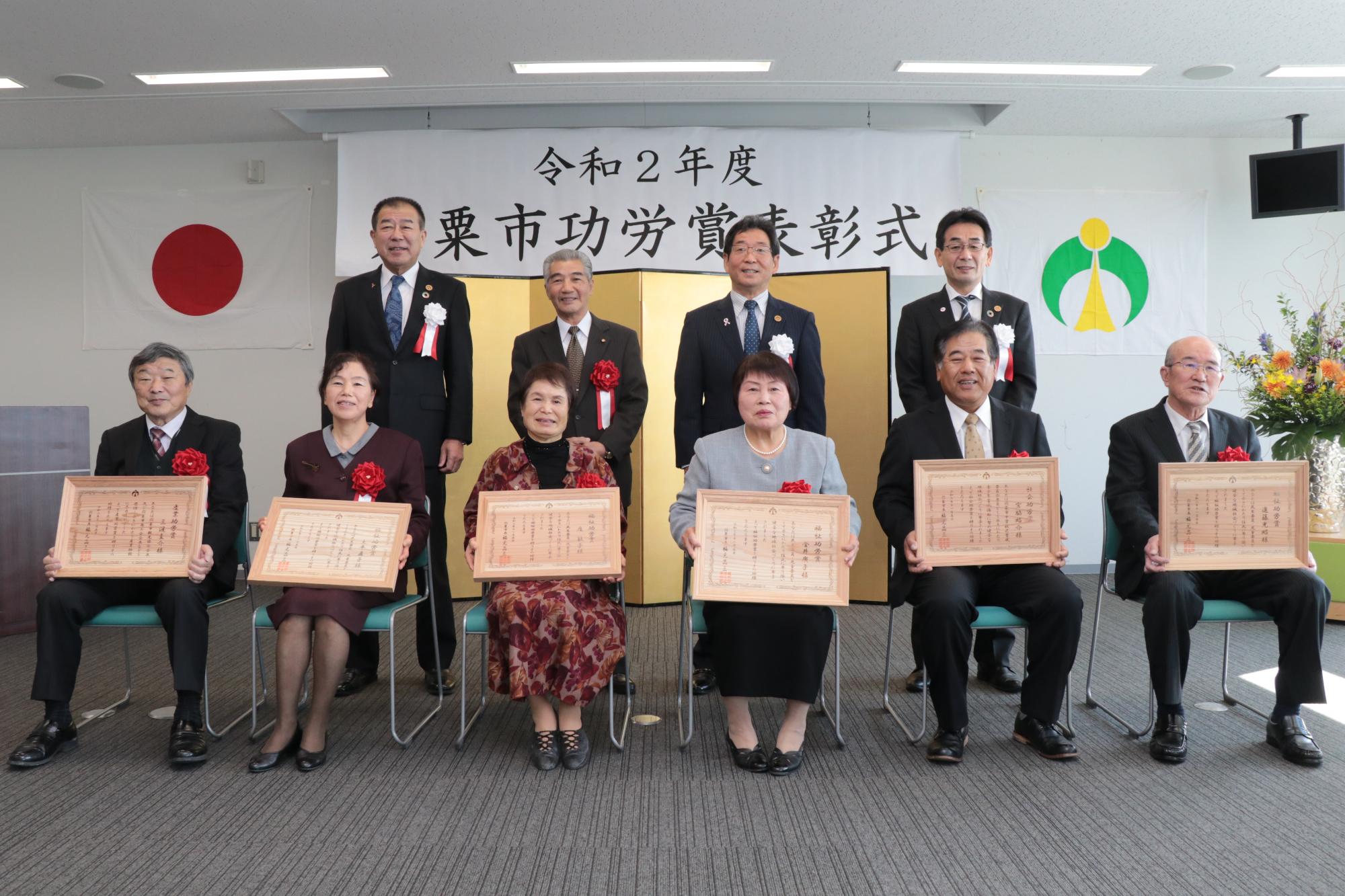 表彰会場で受章者6名の後ろに右から西岡教育長、東議長、福元市長、中村副市長が並ぶ写真