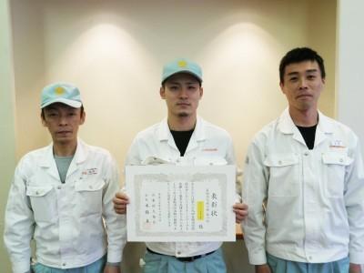 白い作業着を着た3人が賞状を手にしている記念写真