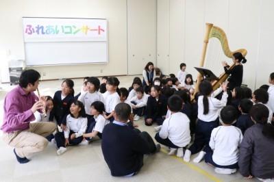 フルートを吹く男性とハープを弾く女性が離れて演奏しそれぞれを児童たちが囲んで座り演奏を見入っている写真