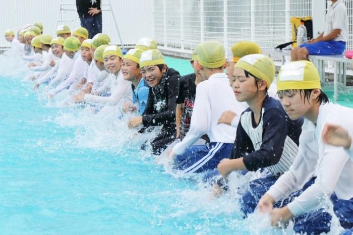 黄色い水泳帽をかぶり服を着てプールの縁で水を自分にかけている男女小学生達の写真