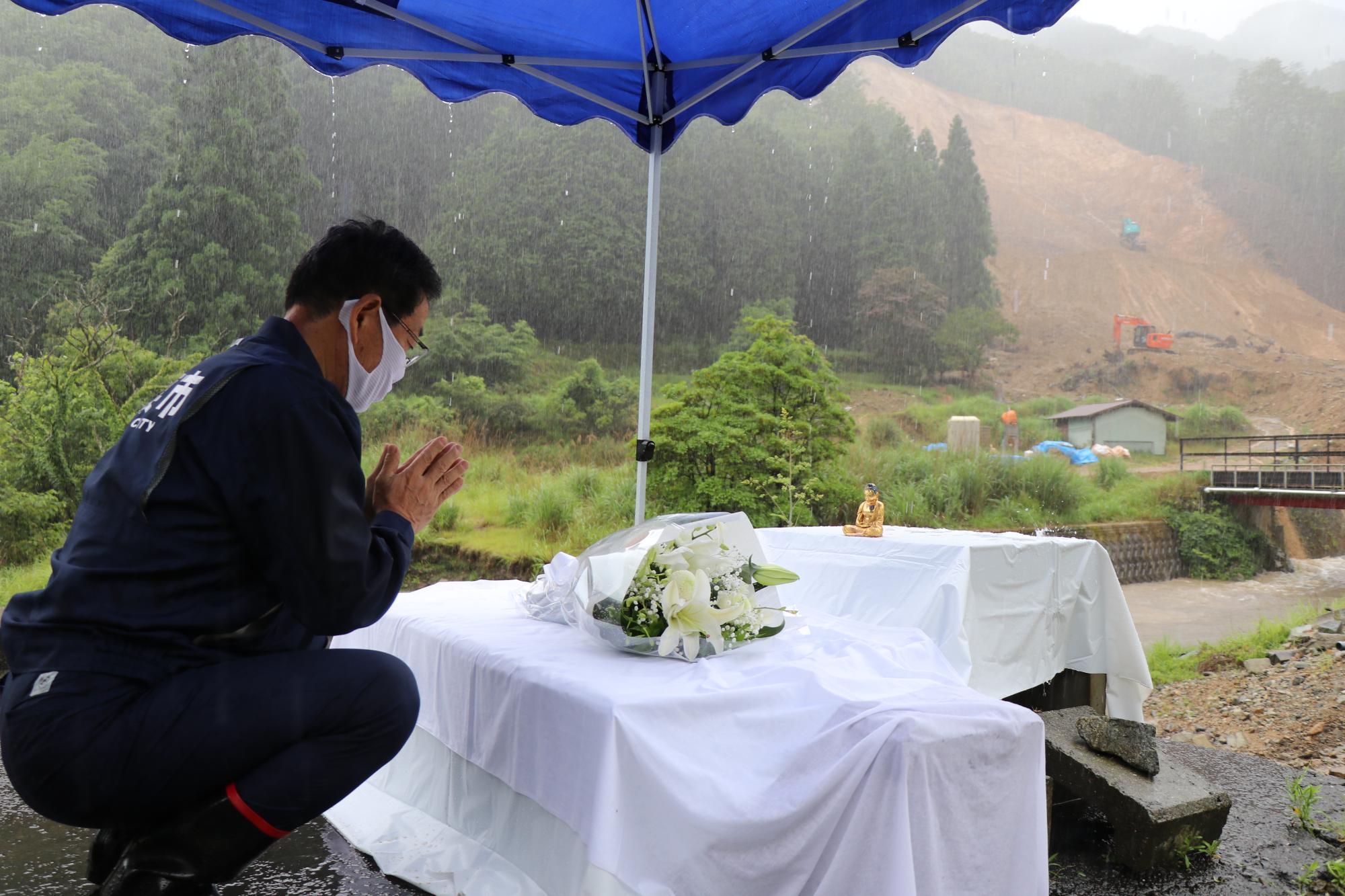 雨の中復旧工事の現場を前に花を供え手を合わせる市長の写真