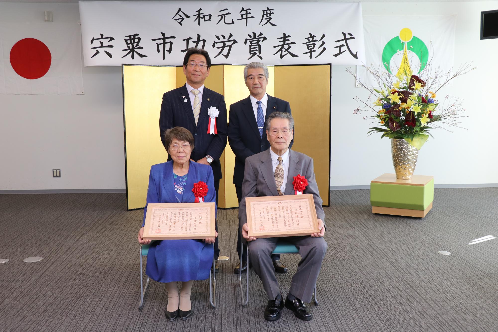 賞状を持った功労賞受賞者2名と市長と議長が金屏風の前に並んでいる写真