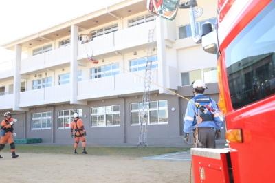 校舎の3階にはしごがかけてあり、近くに2名のオレンジ色の作業着を着た消防隊員が作業をしている写真