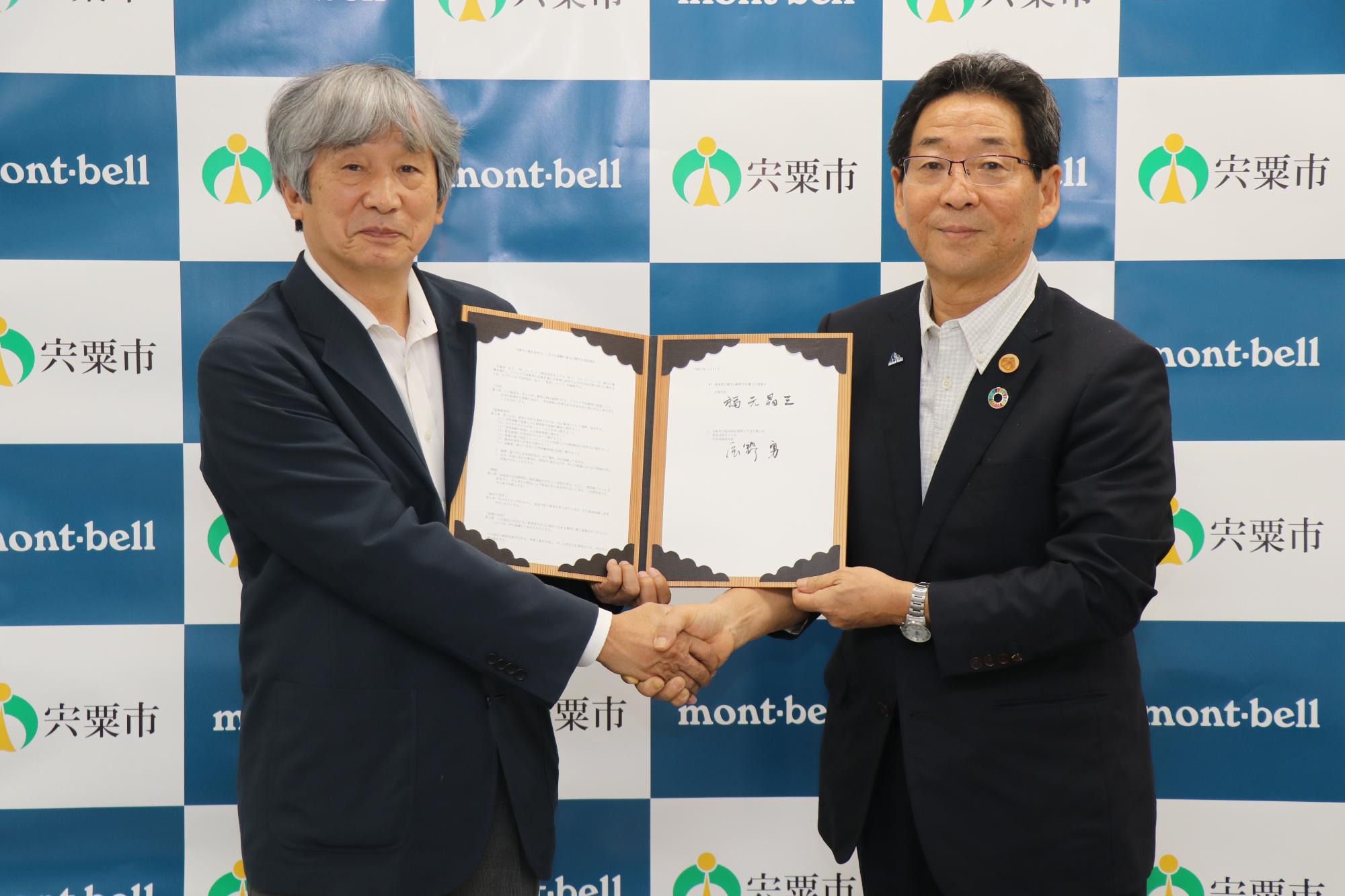 協定書を手に株式会社モンベル辰野会長と握手を交わす市長の写真