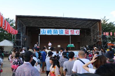 山崎納涼夏祭りのステージで福引をひく福元市長とそれを見守る観客らの写真