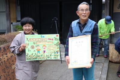 園児たちから送られた園児たちが書いた絵と賞状をもつ小田さん夫妻の写真