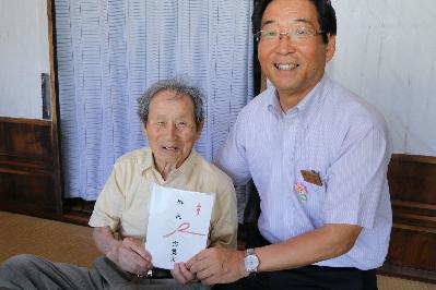 長寿の祝い品を持って笑う最高齢夫妻の森脇高一さんと福元市長の写真