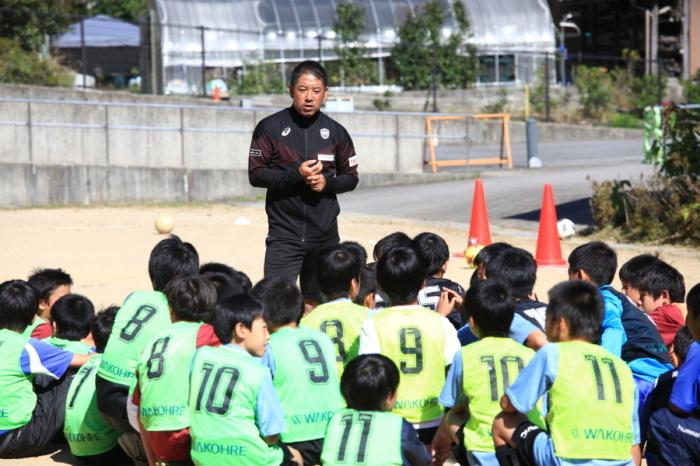 黒いジャージを着たヴィッセル神戸の指導員がしゃがんでいるゼッケンを着た小学生に話をしている写真