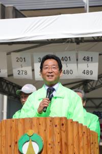 屋外の演壇で明るい蛍光グリーンのジャケットを着た市長がスピーチをしてる写真