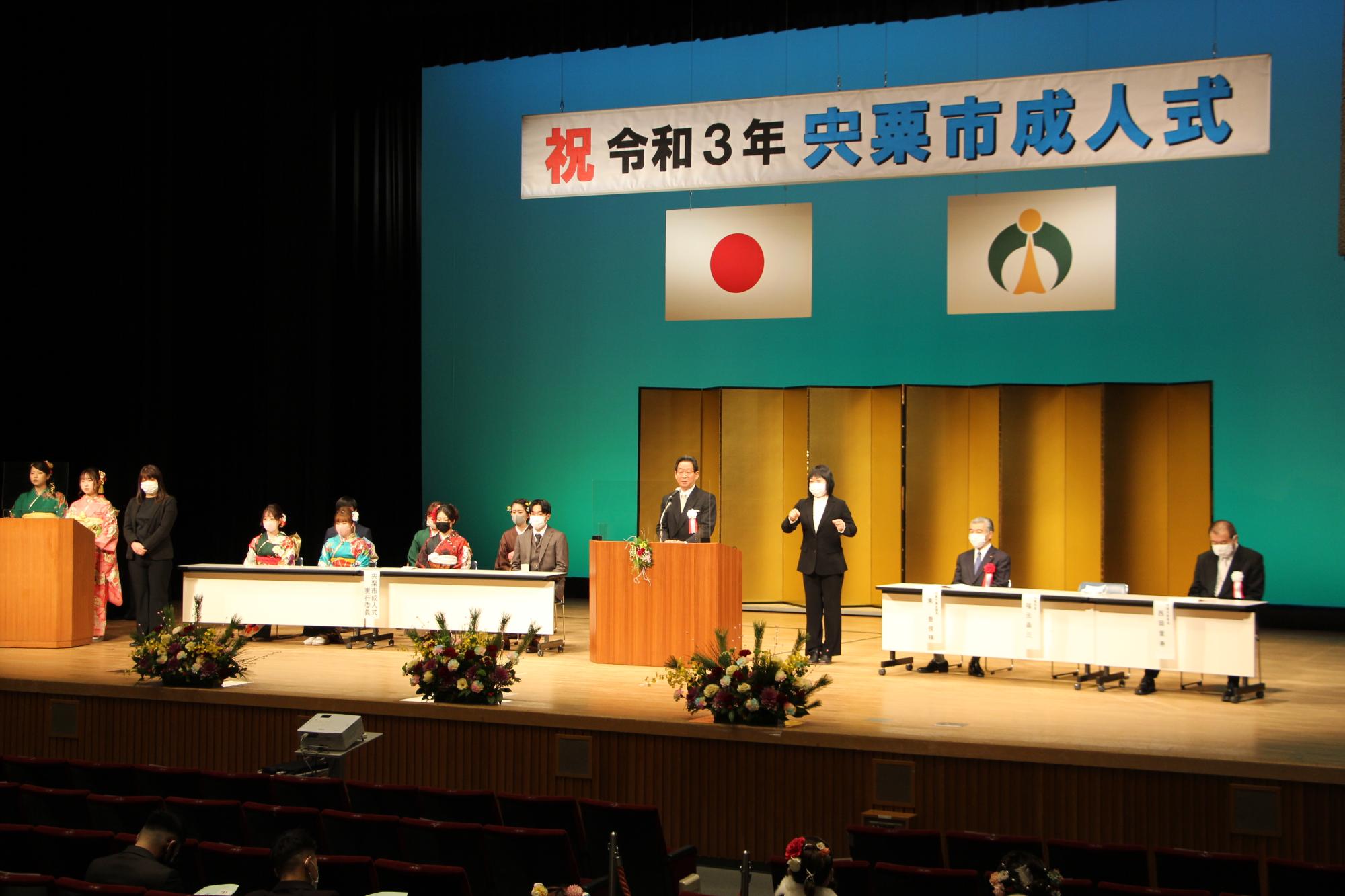 壇上の左側から新成人、中央にあいさつする福元市長、手話通訳を挟んで東議長、西岡教育長の写真。