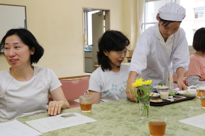 テーブルについている女性の前にランチを配膳する割烹着姿の高校生の写真