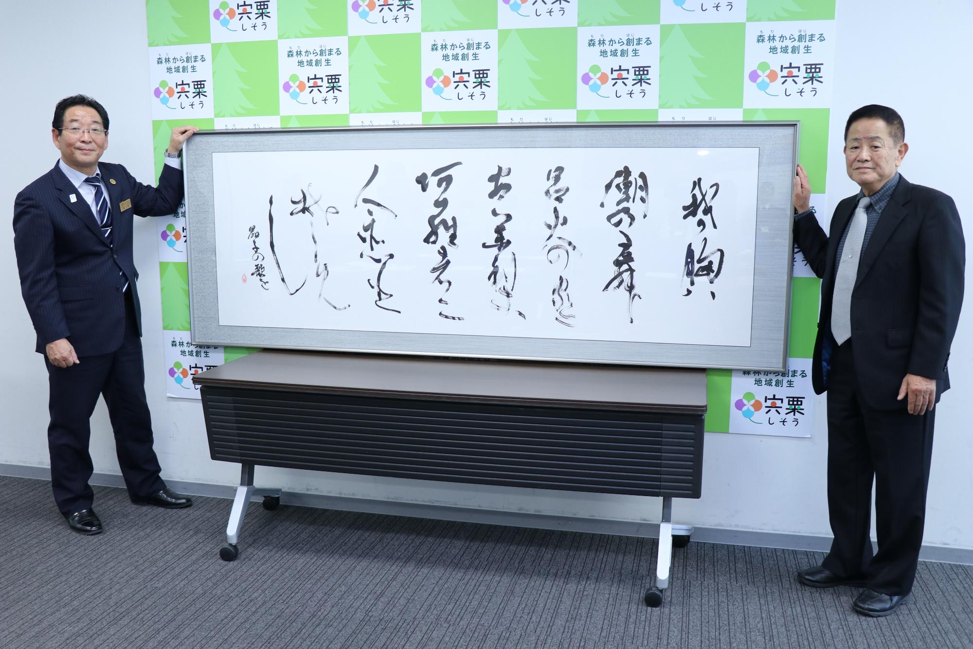 額に入った横に長い大きな紙に書かれた山部一翠氏の作品と市長と製作者が写る記念写真