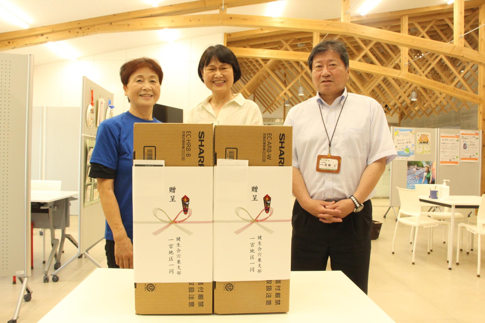 寄贈された掃除機2台を前に、健生会の岡田さん（左）と下村さん（中）が田路局長と並んで立っている写真
