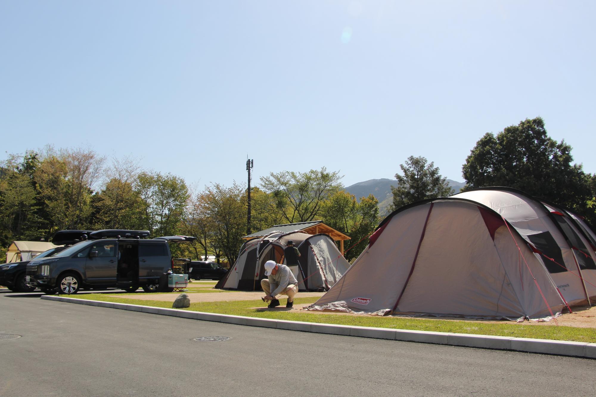 青空のもと、キャンプ場利用者がテントのペグを地面に打ち込んでいる写真