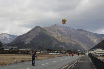 山脈を背景に、道路横の歩道から、1人の男性が凧を飛ばしている様子を後ろから写した写真