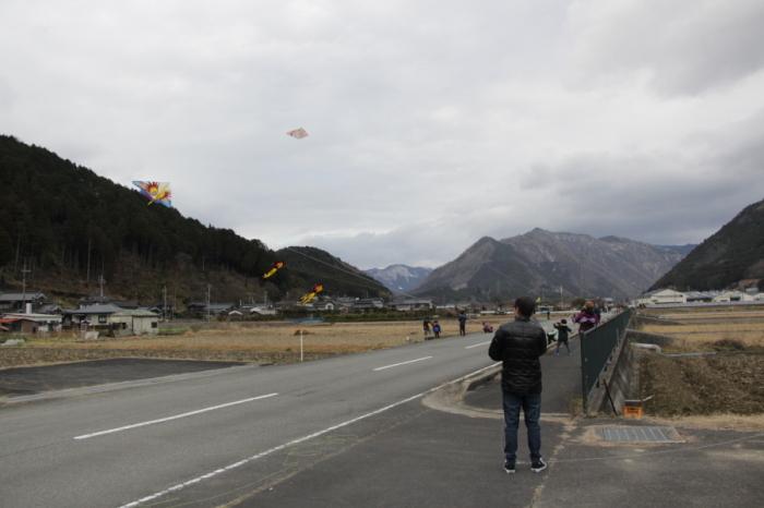山脈を背景に、道路横の歩道から、約6人の人が凧やゲイラカイトを飛ばしている様子を写した写真