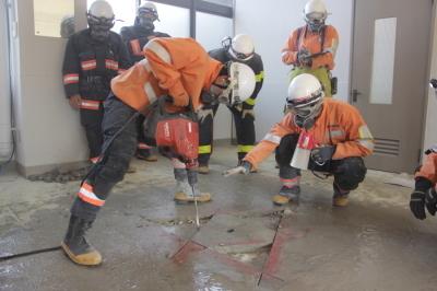 ヘルメットとガスマスクを装着した消防組合員が、床に空けられた三角形の亀裂に、電動ハンマーを打ち立てている様子を、少し離れた場所から撮影した写真