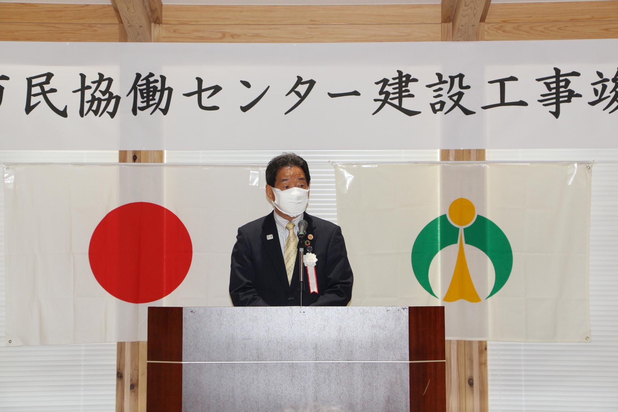 マスクをした福元市長が演台で竣工式の挨拶をしている写真