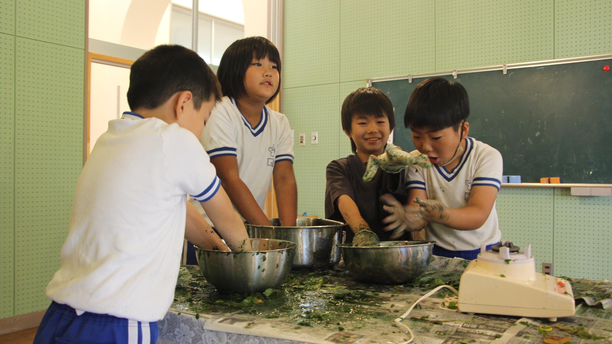 児童らがボウルに入った藍染め液に巾着を漬け込み。浸透させるために揉んでいる写真