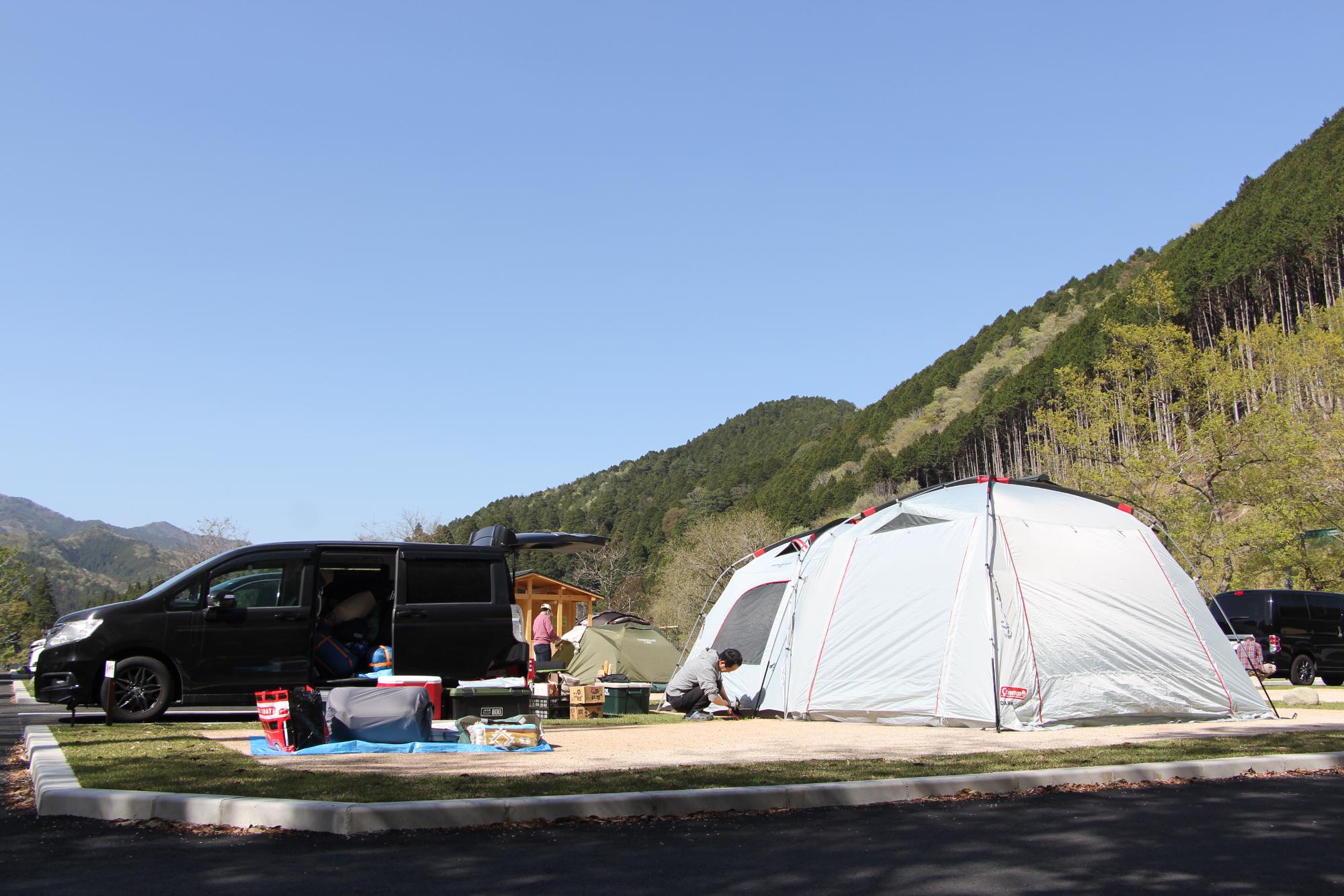 キャンプ場利用者がキャンプ用品を区画内に広げ、テントのペグを地面に打ち込んでいる写真