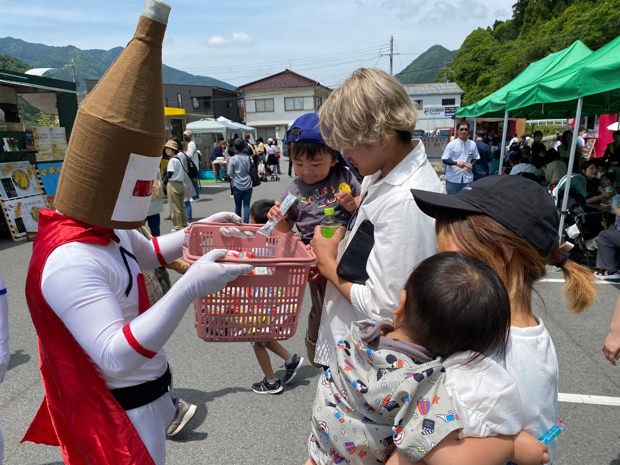 いちにぴあ公認キャラクター「日本酒マン」から来場した親子連れがお菓子を受け取っている写真
