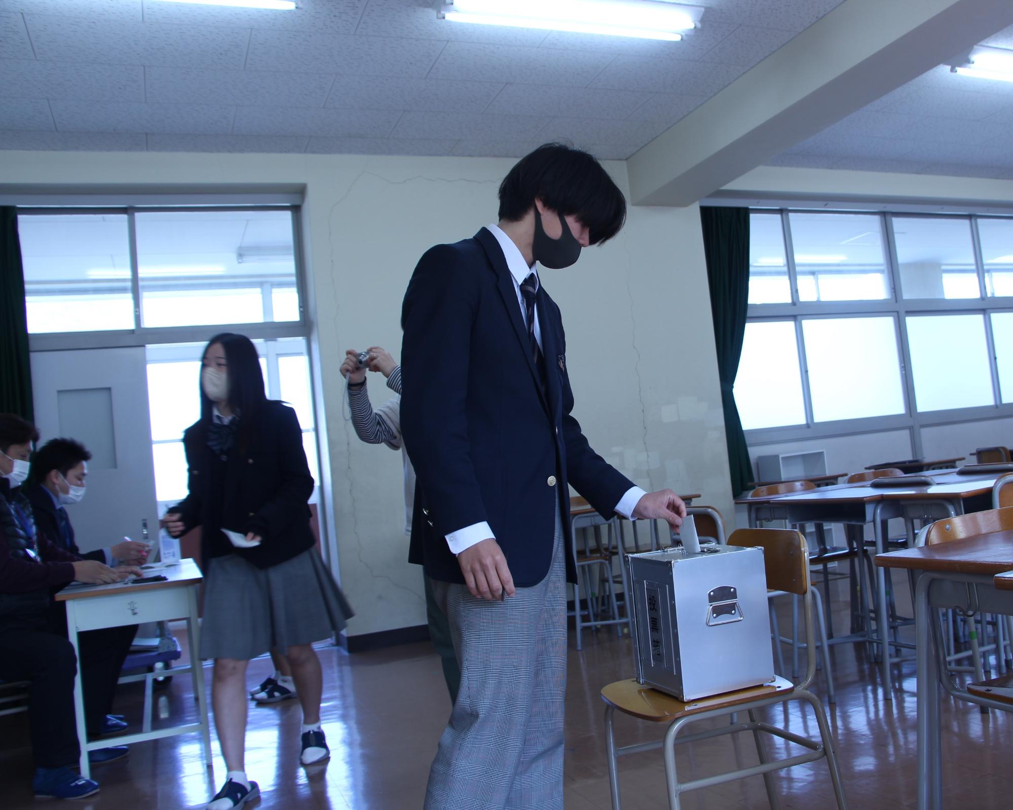 教室内に設置した投票箱へ候補者名を記入した投票用紙を投函する高校生の写真