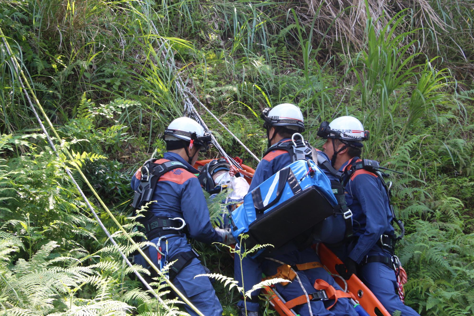 山の急斜面をロープで降りた3人の隊員が山道を滑落した人を想定し、担架に乗せて救助している写真