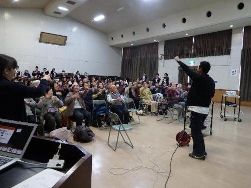 理学療法士の小林憲人さんが左手を上げて「転倒予防」について講演を高齢者にしている写真