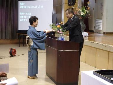 黒いスーツを着た男性が、青い着物を着た女性に賞状を渡している卒業式、修了式の様子