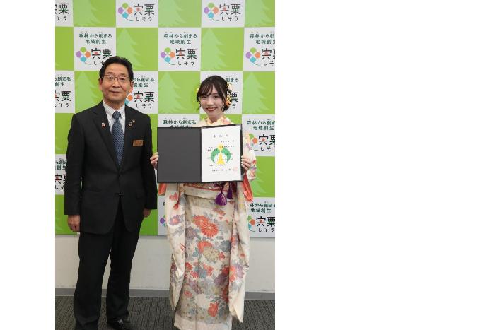 mayu（まゆ）さんが福元晶三市長から手渡された委嘱状を胸の前で広げた状態で、市長と並んで立っている写真