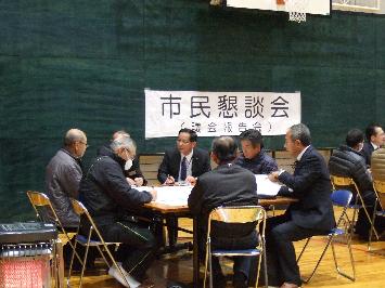体育館で机を囲むスーツを着た8人の男性が意見を交わしている、懇談会の写真