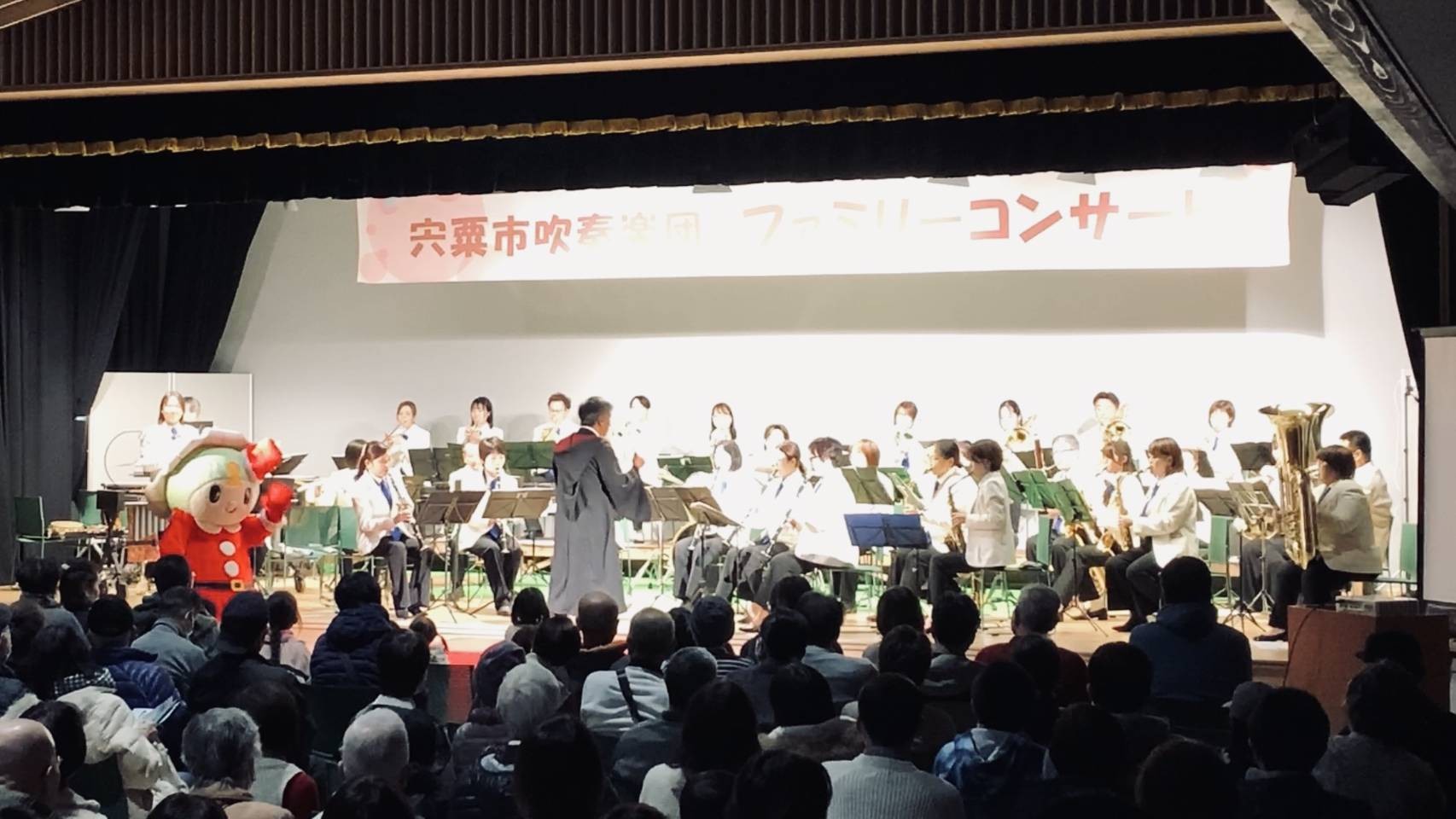 はがてらす大ホールのステージで、宍粟市吹奏楽団のメンバーが隊形に並んで座り、楽器を演奏している写真