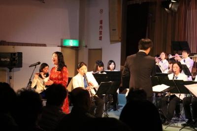 オレンジのドレスを着た中村理恵さんが吹奏楽部の前でびやかな歌声で歌っている写真