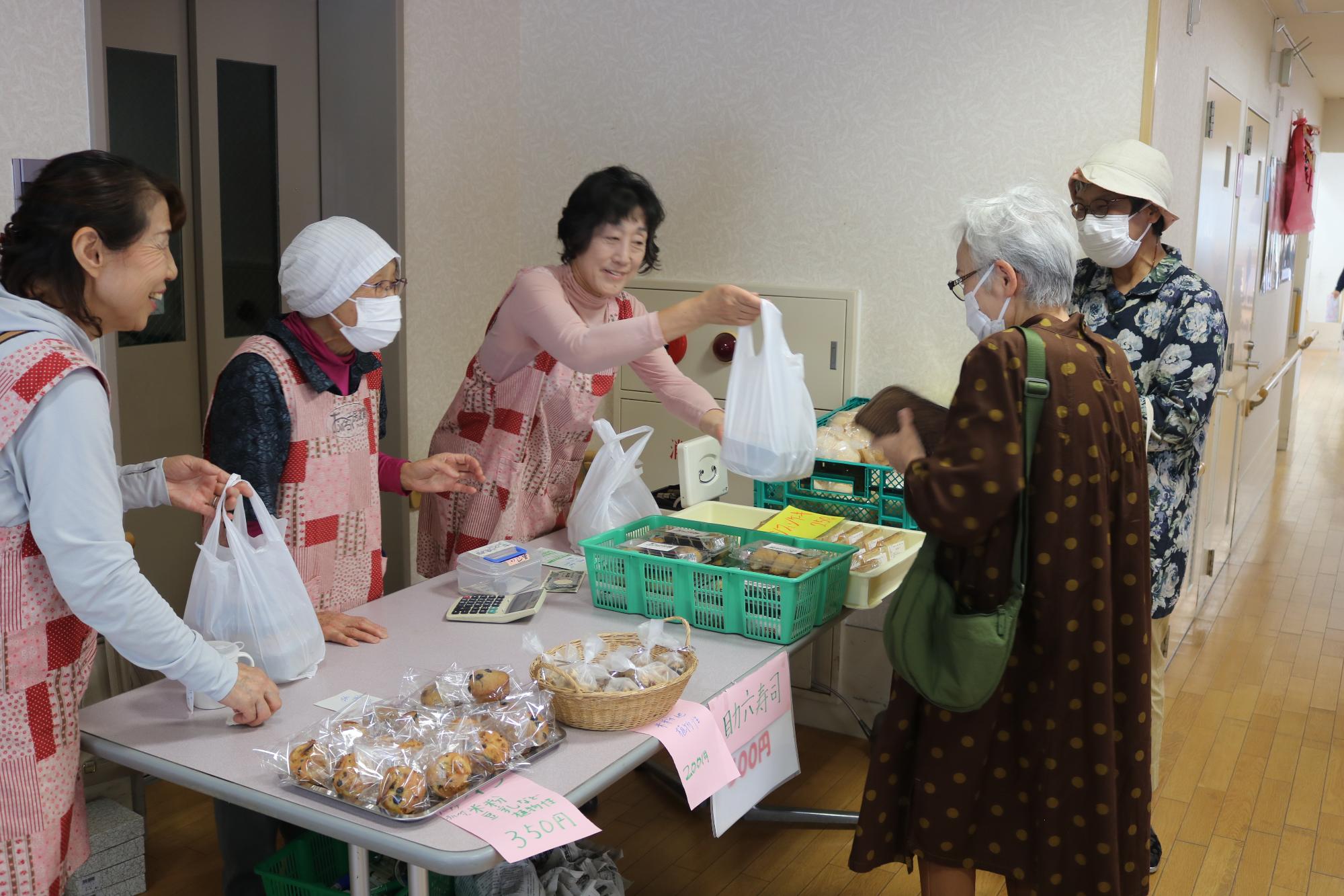 巻き寿司やいなり寿司、洋菓子などを販売するフリーマーケットで来場者が購入した商品を店員から受け取っている写真