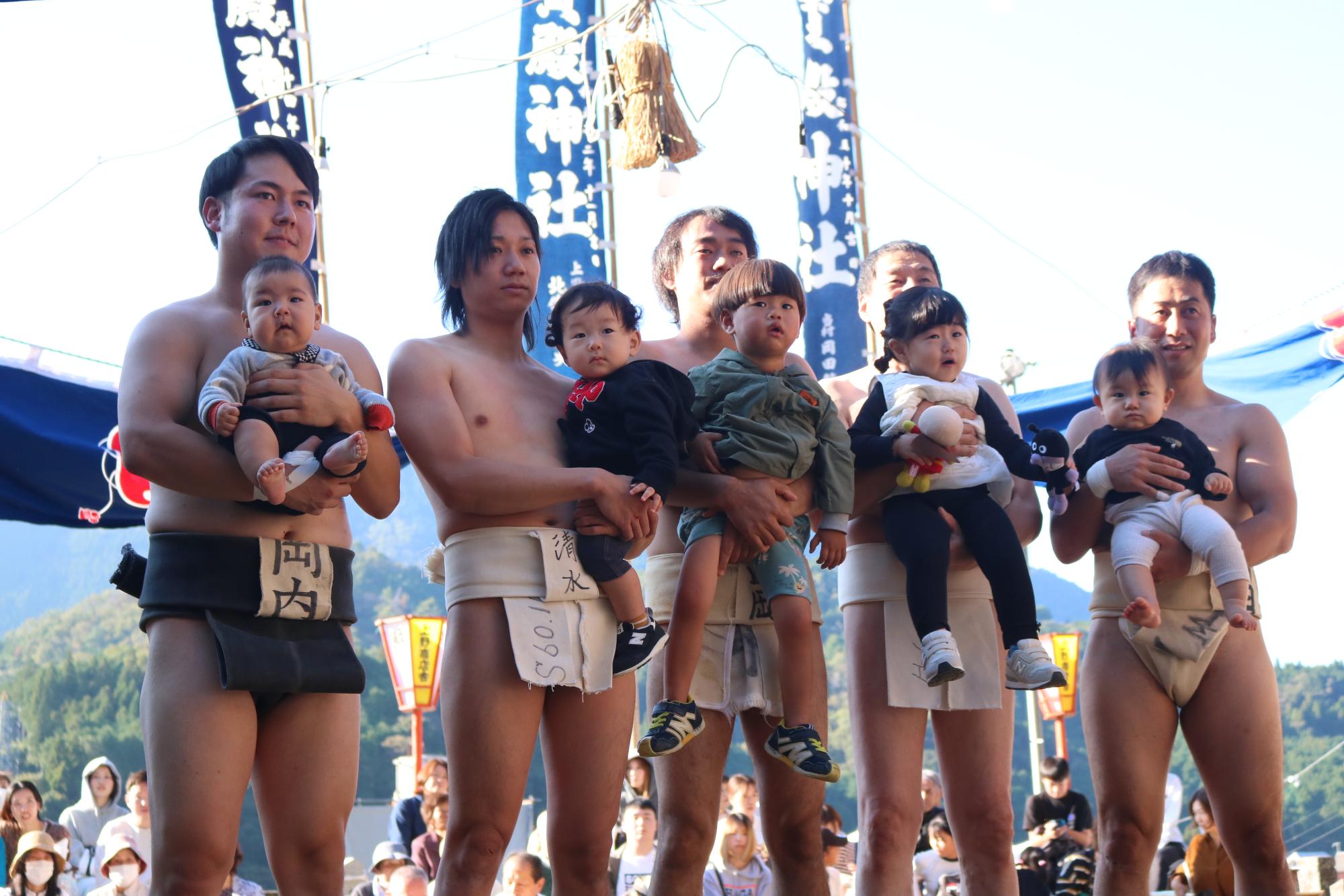 5人の力士が小さな子ども抱えて土俵に立っている写真