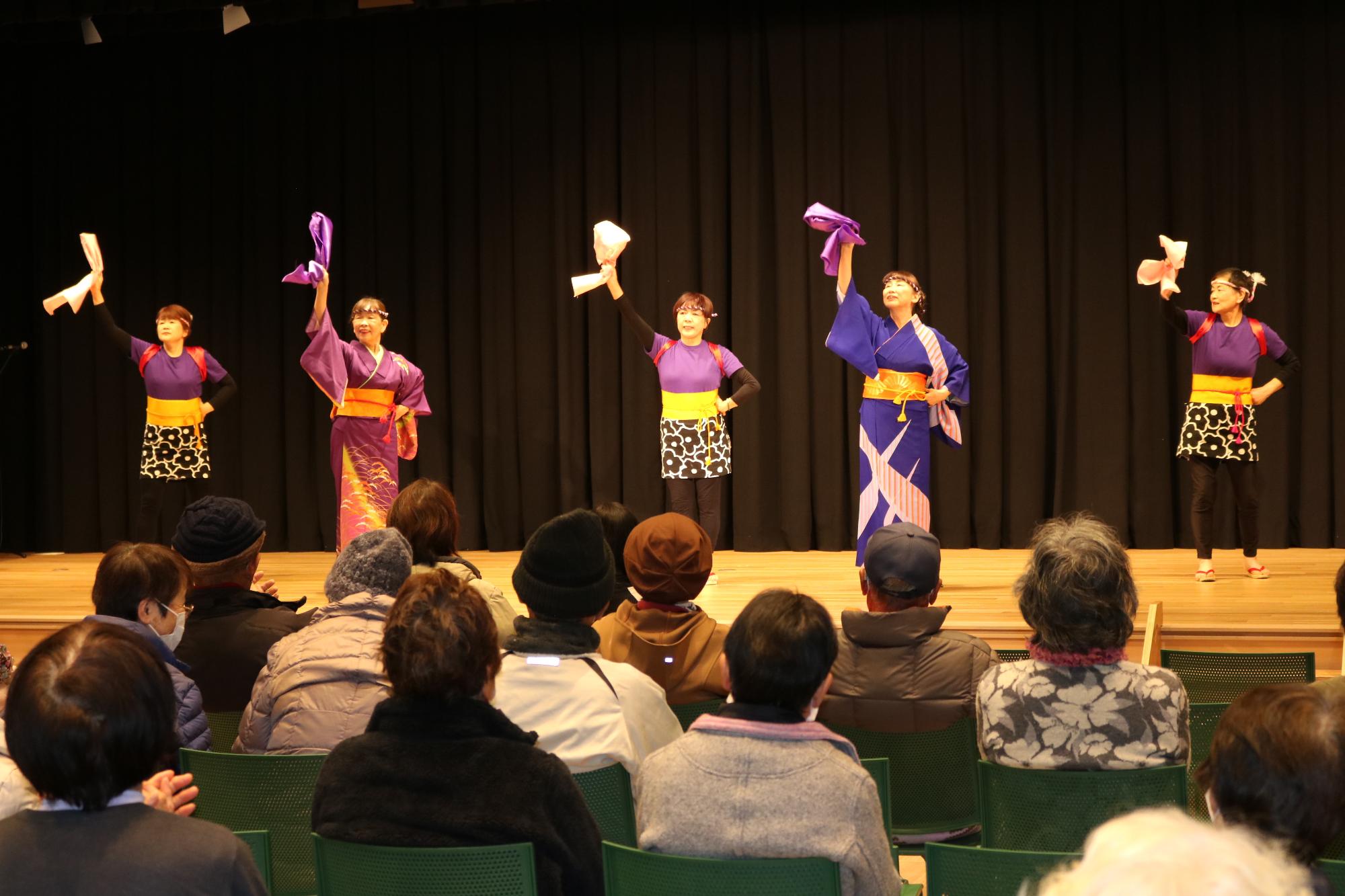 田舎の家マミーズのメンバーが、はがてらすのステージで歌と踊りを披露している写真