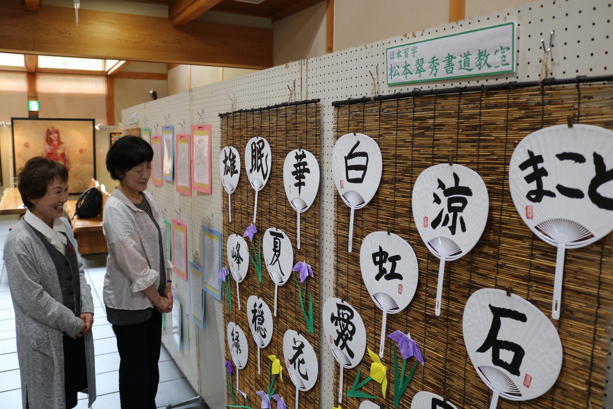 折り紙の花と白い面に墨で字が書かれた団扇が簾の上に並べて貼り付けてある展示作品を女性ふたりが見ている写真